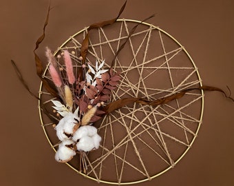 May include: A brown, circular, woven macrame wall hanging with a gold metal hoop. The macrame is decorated with dried flowers and cotton bolls in shades of pink, white, and brown.