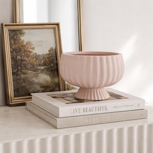 Op de afbeelding: Een lichtroze, geribbelde keramische kom met een voetstuk. De kom staat op twee boeken met de titels "LIVE BEAUTIFUL" en "INTERIORS". Op de achtergrond staat een ingelijst landschapsschilderij.