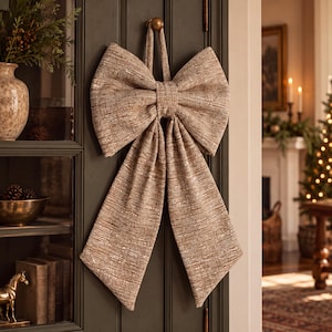 May include: A large decorative bow made of textured, light brown fabric with a subtle metallic sheen. The bow features two wide loops at the top and two long, hanging tails that taper toward the bottom, suspended by a fabric loop.