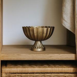 May include: A gold-coloured, pedestal bowl with a fluted design. The bowl has a scalloped rim and sits on a decorative base. It is displayed on a wooden shelf, suggesting a decorative or display item.