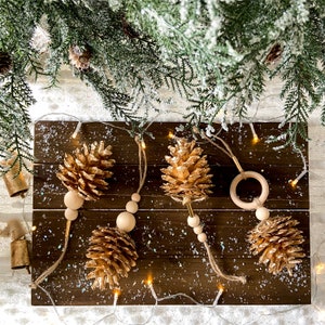 May include: Gold-painted pine cone decorations with wooden beads and twine hanging on a dark brown wooden surface. The decorations are arranged with fairy lights and faux evergreen branches, creating a festive holiday display.