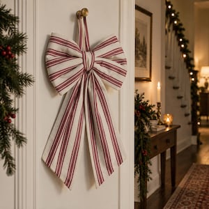 May include: A large decorative bow made of off-white fabric with vertical dark red stripes. The bow features two wide loops and two long, hanging tails, suspended from a small brass-colored hook mounted on a white wall surface.