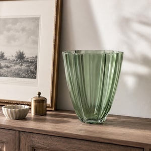 Op de afbeelding: Een grote, saliegroene glazen vaas met verticale geribbelde details. De vaas heeft een uitlopende vorm en staat op een houten oppervlak. Een kleine, ronde, goudkleurige container met een deksel en een kleine, witte, geschulpte kom staan ook op het oppervlak.