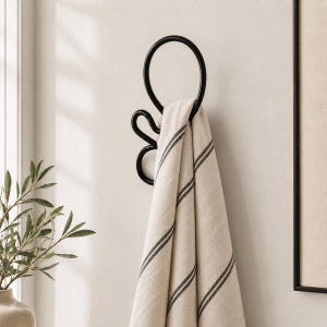 May include: A black metal towel ring with a circular design and a decorative flourish. A beige towel with dark grey stripes hangs from the ring. The towel is made of a soft, textured fabric. The design is minimalist and modern.