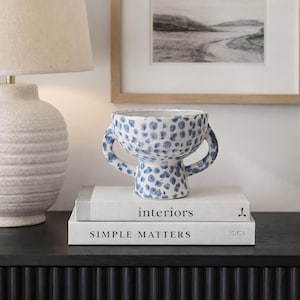 Op de afbeelding: Een keramische vaas met een blauw stippenpatroon en twee handvatten staat op twee boeken. De boeken heten "interiors" en "SIMPLE MATTERS". Een lamp en een ingelijst kunstwerk staan op de achtergrond.