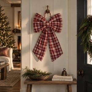 May include: A large decorative bow made of red and white plaid fabric hanging from a brass hook. The bow features two wide loops and two long, tapered tails with a classic checkered pattern, designed for seasonal wall or door ornamentation.