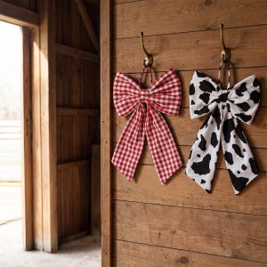 May include: Two decorative bows hanging on a wooden wall. One bow features a red and white gingham pattern, while the other has a black and white cow print. Both are suspended from brass hooks.