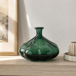 Könnte beinhalten: Eine dunkelgrüne Glasvase mit einem bauchigen, gerippten Körper und einem schmalen Hals. Die Vase steht auf einer hellen Holzoberfläche, mit einem gerahmten Bild und einem kleinen dekorativen Gegenstand im Hintergrund.
