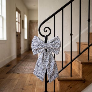 May include: A large fabric bow with a blue floral pattern hangs from a black metal stair railing. The bow is made of a light-colored fabric with a delicate blue floral print. The bow is displayed on a wooden staircase.
