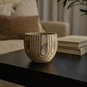 May include: A small, round, beige decorative bowl with a textured, carved wood-grain pattern. The bowl is on a dark surface. The background shows a sofa and books.