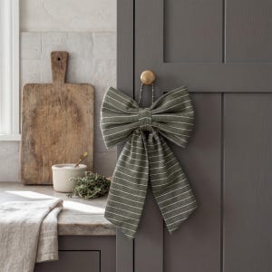 May include: A large, olive green fabric bow with white stripes hangs from a gold knob on a gray cabinet door. The bow is a decorative accent, possibly for a kitchen or dining area. A wooden cutting board and other kitchen items are in the background.