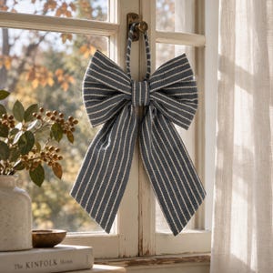 May include: A decorative fabric bow featuring a slate blue background with thin, vertical white pinstripes. The bow has a central knot and two long, hanging tails, with a matching fabric loop at the top for hanging on a window latch.