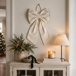 May include: A large, cream-coloured fabric bow with a subtle gold polka dot pattern hangs on a wall. The bow features two wide top loops and two long, pointed tails. It is constructed from a textured textile material and is suspended by a small loop.