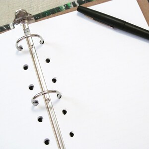 May include: An open white notebook with a silver binder and a black pen resting on the blank pages. The notebook is on a patterned surface.