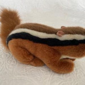 May include: A plush chipmunk toy with a fluffy, reddish-brown tail and body. The chipmunk has a dark brown stripe bordered by white stripes across its back. The toy is lying on a white, textured surface.
