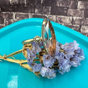 May include: A silver ring with a large, clear, elongated gemstone. The ring is set on a bed of dried blue flowers. The ring's band features a small, sunburst-shaped design with a pearl in the center. The background is a blue surface and a brick wall.