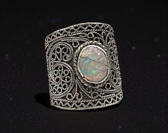Könnte beinhalten: Ein silberner Ring mit einem ovalen Opalstein. Der Ring weist aufwendige Filigranarbeiten um den Stein und das Band auf. Der Opal hat ein rissiges Aussehen und zeigt schillernde Farben. Der Ring ist vor einem dunklen Hintergrund platziert.
