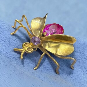 May include: A gold-toned insect brooch with a large pink gemstone and a smaller purple gemstone. The brooch has detailed wings and legs, and is set against a blue background.