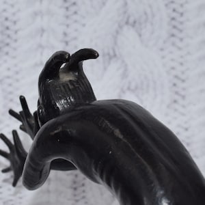 Könnte beinhalten: Eine schwarze Skulptur einer Kreatur mit ausgestreckten Armen und einem Kopf mit zwei Hörnern. Die Skulptur hat eine glatte, glänzende Oberfläche und befindet sich vor einem weißen, strukturierten Hintergrund.