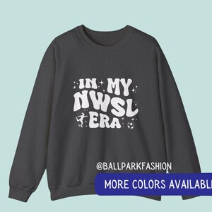 Op de afbeelding: Donkergrijze sweatshirt met de witte tekst "IN MY NWSL ERA", een silhouet van een voetballer en een voetbalafbeelding. De sweatshirt heeft ook de tekst "@BALLPARKFASHION" en "MORE COLORS AVAILABLE" in een blauwe banner.
