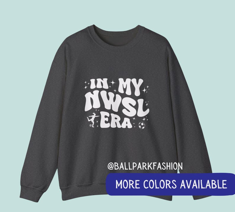 Op de afbeelding: Donkergrijze sweatshirt met de witte tekst "IN MY NWSL ERA", een silhouet van een voetballer en een voetbalafbeelding. De sweatshirt heeft ook de tekst "@BALLPARKFASHION" en "MORE COLORS AVAILABLE" in een blauwe banner.