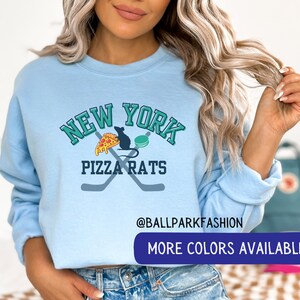 May include: A light blue sweatshirt with a graphic design that says "New York Pizza Rats" with a rat eating a slice of pizza and a hockey puck. The text "@ballparkfashion" and "More Colors Available" are also on the sweatshirt.