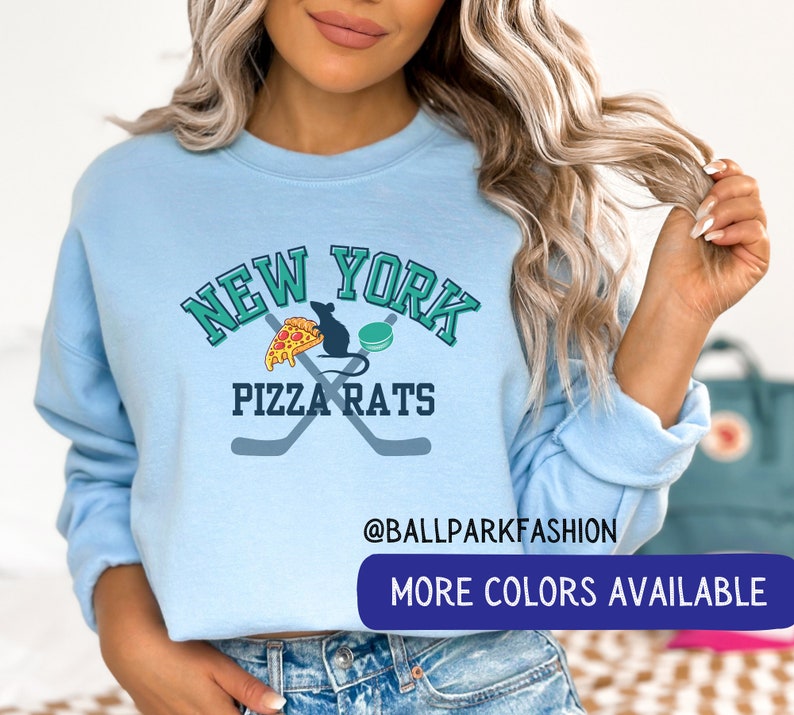 May include: A light blue sweatshirt with a graphic design that says "New York Pizza Rats" with a rat eating a slice of pizza and a hockey puck. The text "@ballparkfashion" and "More Colors Available" are also on the sweatshirt.