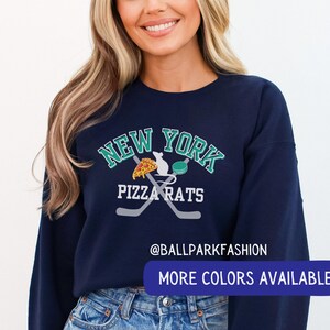 May include: A navy blue sweatshirt with a white graphic of a hockey stick and a rat holding a slice of pizza. The graphic also includes the text "New York Pizza Rats" and "@ballparkfashion". The text "More Colours Available" is at the bottom of the image.