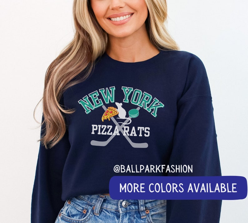 May include: A navy blue sweatshirt with a white graphic of a hockey stick and a rat holding a slice of pizza. The graphic also includes the text "New York Pizza Rats" and "@ballparkfashion". The text "More Colours Available" is at the bottom of the image.