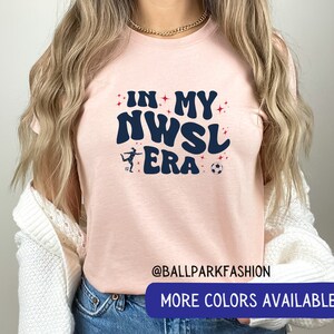 Pu&ograve; includere: Maglietta rosa con il testo "In My NWSL Era" in un font retr&ograve;. Una silhouette di un calciatore &egrave; presente sulla maglietta. Sono visibili anche i testi "@ballparkfashion" e "Altri colori disponibili".