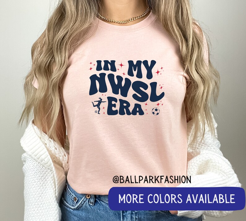Pu&ograve; includere: Maglietta rosa con il testo "In My NWSL Era" in un font retr&ograve;. Una silhouette di un calciatore &egrave; presente sulla maglietta. Sono visibili anche i testi "@ballparkfashion" e "Altri colori disponibili".