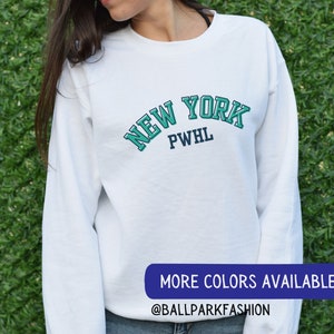 May include: White long-sleeved sweatshirt with a teal blue and navy blue graphic that says "NEW YORK PWHL". The text is in a vintage collegiate style font. The sweatshirt is being modelled by a person. The text "MORE COLORS AVAILABLE @BALLPARKFASHION" is at the bottom of the image.
