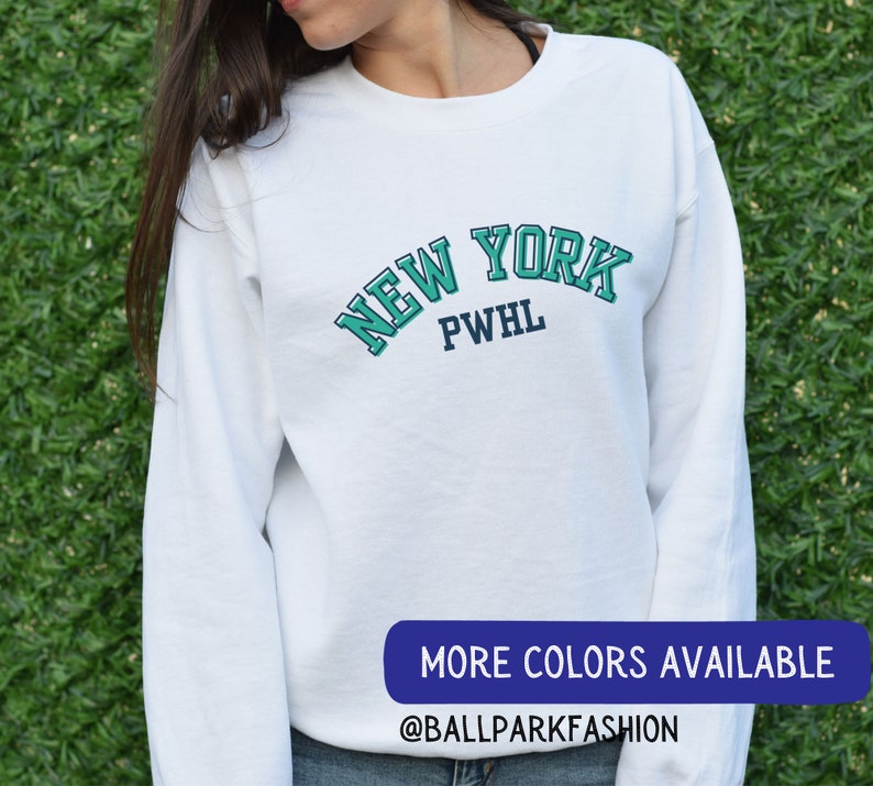 May include: White long-sleeved sweatshirt with a teal blue and navy blue graphic that says "NEW YORK PWHL". The text is in a vintage collegiate style font. The sweatshirt is being modelled by a person. The text "MORE COLORS AVAILABLE @BALLPARKFASHION" is at the bottom of the image.