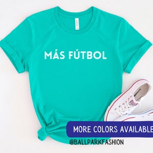Puede incluir: Una camiseta turquesa con el texto "M&Aacute;S F&Uacute;TBOL" en blanco. La camiseta est&aacute; anudada en la parte inferior. El texto "MORE COLORS AVAILABLE @BALLPARKFASHION" est&aacute; en la parte inferior de la imagen.