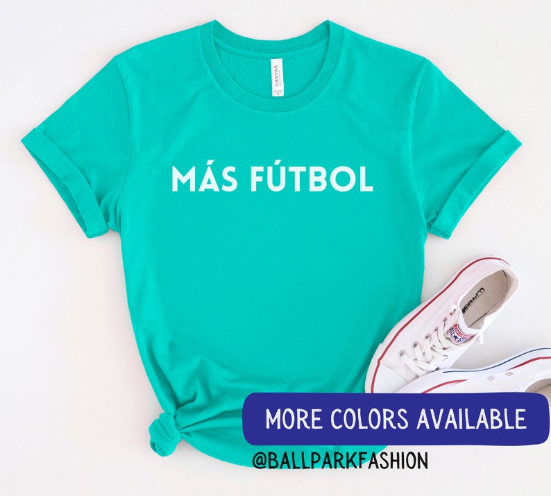 Puede incluir: Una camiseta turquesa con el texto "M&Aacute;S F&Uacute;TBOL" en blanco. La camiseta est&aacute; anudada en la parte inferior. El texto "MORE COLORS AVAILABLE @BALLPARKFASHION" est&aacute; en la parte inferior de la imagen.