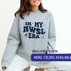 Op de afbeelding: Een lichtgrijs sweatshirt met de tekst "IN MY NWSL ERA" in marineblauw, rood en wit. Het ontwerp bevat een silhouet van een voetballer en een voetbal. De sweatshirt heeft een ronde hals en lange mouwen.