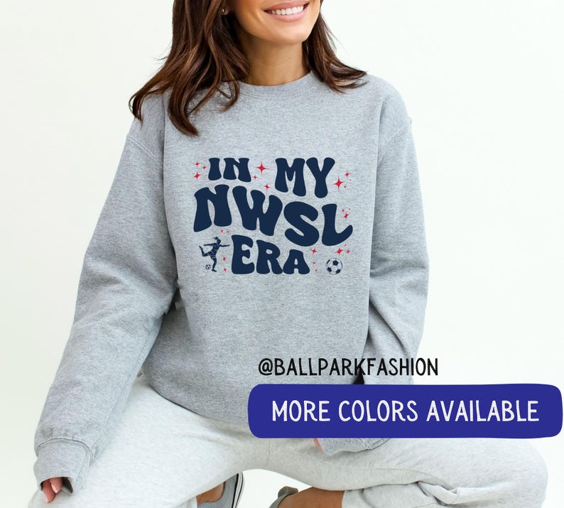 Op de afbeelding: Een lichtgrijs sweatshirt met de tekst "IN MY NWSL ERA" in marineblauw, rood en wit. Het ontwerp bevat een silhouet van een voetballer en een voetbal. De sweatshirt heeft een ronde hals en lange mouwen.