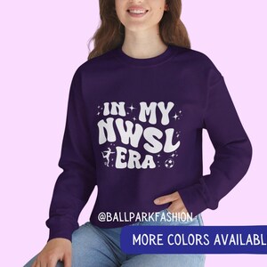 Op de afbeelding: Een donkerpaarse sweatshirt met de witte tekst "IN MY NWSL ERA", een silhouet van een voetballer en een voetbal. De sweatshirt heeft ook een blauwe banner met de tekst "MORE COLORS AVAILABLE".