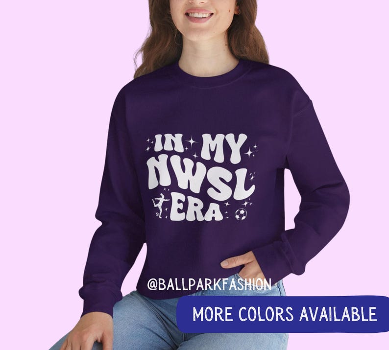 Op de afbeelding: Een donkerpaarse sweatshirt met de witte tekst "IN MY NWSL ERA", een silhouet van een voetballer en een voetbal. De sweatshirt heeft ook een blauwe banner met de tekst "MORE COLORS AVAILABLE".