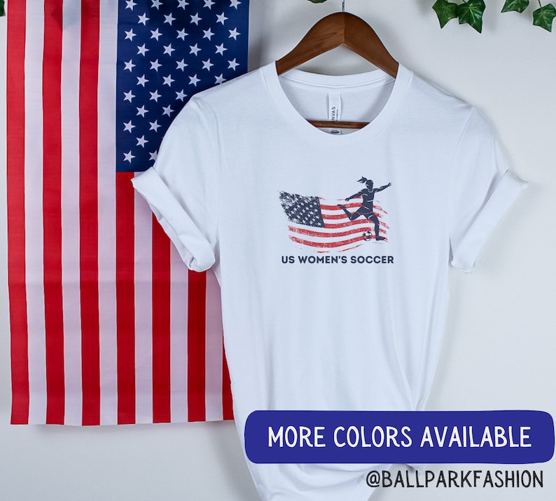 Puede incluir: Camiseta blanca con un gr&aacute;fico de bandera estadounidense desgastado y una silueta de un jugador de f&uacute;tbol pateando una pelota. El texto "US Women's Soccer" est&aacute; impreso debajo del gr&aacute;fico.
