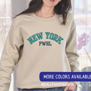 May include: A beige sweatshirt with a teal blue and navy blue graphic that says "NEW YORK PWHL" on the front.