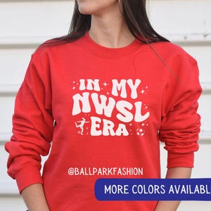 Op de afbeelding: Een rode sweatshirt met de tekst "IN MY NWSL ERA" in het wit, samen met een voetbal en een afbeelding van een voetballer. De sweatshirt heeft ook de tekst "@BALLPARKFASHION" en een knop met de tekst "MEER KLEUREN BESCHIKBAAR".