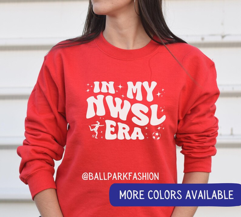 Op de afbeelding: Een rode sweatshirt met de tekst "IN MY NWSL ERA" in het wit, samen met een voetbal en een afbeelding van een voetballer. De sweatshirt heeft ook de tekst "@BALLPARKFASHION" en een knop met de tekst "MEER KLEUREN BESCHIKBAAR".