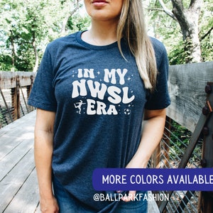Pu&ograve; includere: Una maglietta blu navy con il testo "In My NWSL Era" in bianco con una grafica stilizzata di un calciatore e un pallone da calcio. Il testo &egrave; in un font retr&ograve;. La maglietta &egrave; indossata da una persona in piedi su un ponte di legno. Il testo "More Colors Available @BallparkFashion" &egrave; nella parte inferiore dell'immagine.