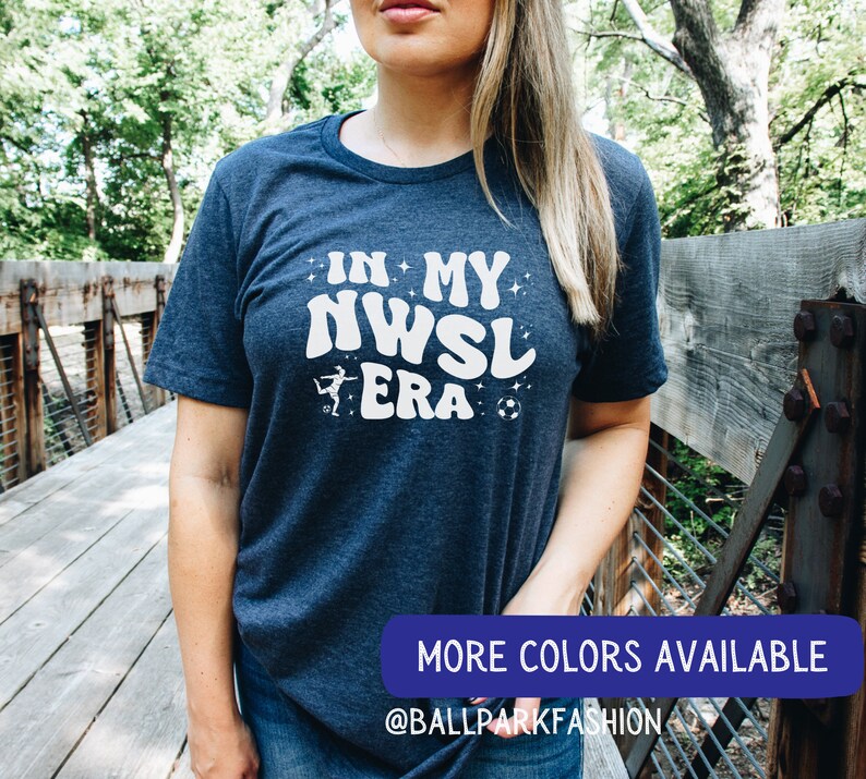 Pu&ograve; includere: Una maglietta blu navy con il testo "In My NWSL Era" in bianco con una grafica stilizzata di un calciatore e un pallone da calcio. Il testo &egrave; in un font retr&ograve;. La maglietta &egrave; indossata da una persona in piedi su un ponte di legno. Il testo "More Colors Available @BallparkFashion" &egrave; nella parte inferiore dell'immagine.