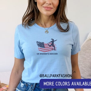 Puede incluir: Camiseta azul claro con un gr&aacute;fico de una jugadora de f&uacute;tbol femenino pateando un bal&oacute;n de f&uacute;tbol frente a una bandera estadounidense. El texto "US Women's Soccer" est&aacute; debajo del gr&aacute;fico. El texto "@ballparkfashion" est&aacute; en la parte inferior de la camiseta. El texto "M&aacute;s colores disponibles" est&aacute; en la parte inferior de la imagen.