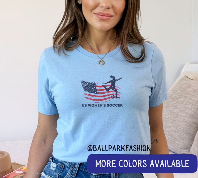 Puede incluir: Camiseta azul claro con un gr&aacute;fico de una jugadora de f&uacute;tbol femenino pateando un bal&oacute;n de f&uacute;tbol frente a una bandera estadounidense. El texto "US Women's Soccer" est&aacute; debajo del gr&aacute;fico. El texto "@ballparkfashion" est&aacute; en la parte inferior de la camiseta. El texto "M&aacute;s colores disponibles" est&aacute; en la parte inferior de la imagen.