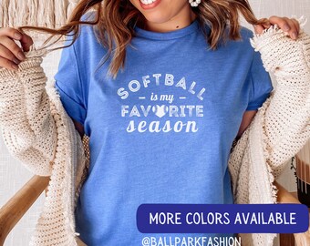 May include: A blue t-shirt with white text that reads "Softball is my favorite season" with a white baseball graphic inside a heart. The text "More Colors Available" and "@ballparkfashion" are printed in white on a blue rectangle at the bottom of the shirt.