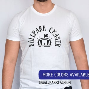 Puede incluir: Camiseta blanca con texto negro que dice "Ballpark Chaser" en semicírculo alrededor de una silueta negra de un estadio de béisbol. El texto "More Colors Available" y "@ballparkfashion" están impresos en negro sobre un rectángulo azul en la parte inferior de la imagen.