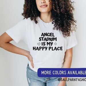 May include: A white t-shirt with black text that reads "Angel Stadium is my happy place" with a heart and baseball graphic. The text is in a handwritten font. The model is wearing the shirt and has long curly brown hair. The text "More colors available @ballparkfashion" is at the bottom of the image.